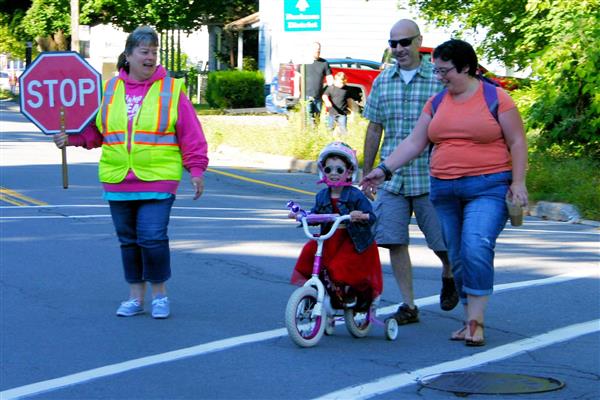 Adults walking a child on a tricycle across the street while a crossing guard helps them pass